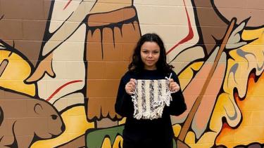 Student stands in front of mural and holds completed Salish weaving
