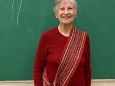 Woman Elder stands in front of green chalkboard wearing red long sleeve and Metis sash