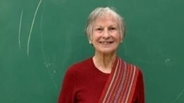 Woman Elder stands in front of green chalkboard wearing red long sleeve and Metis sash