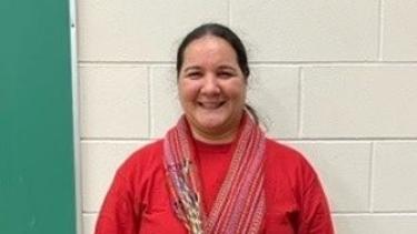 Metis woman stands in front of white brick wall. She wears a red long sleeve and metis sashes.