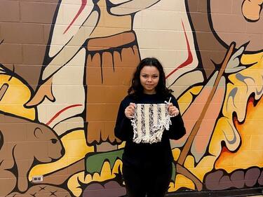 Student stands in front of mural and holds completed Salish weaving