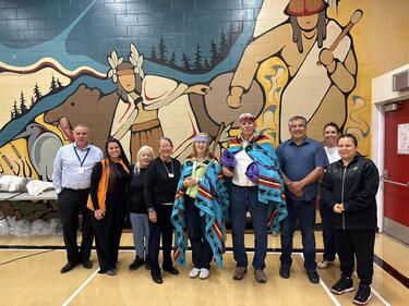 Group of people standing in front of a wall of Indigenous artwork. Two of the people blanketed as an honouring.