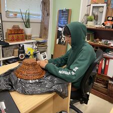Student sitting at desk working on cedar weaving at hat