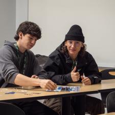 Two male students sitting at a table, one is looking at camera and giving a thumbs up