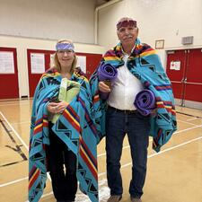 A female and male standing in a gymnasium. Both have been blanketed for an Indigenous Honouring.